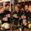 Das Bild Zeigt Eine Entspannte Kaffeepause In Der Feuerwache Bei Der Mehrere Feuerwehrkameraden Und Zwei Frauen Gemeinsam Mit Den Tassen Lachen Und Den Moment Geniessen