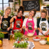 Vier Frauen Beim Gemeinsamen Kochen In Einer Modernen Kueche Jeweils Mit Einer „Chefin Der Pfanne Kuechenschuerze In Schwarz Rot Pink Und Weiss Mit Lustigem Pfannenmotiv